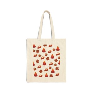Könnte beinhalten: Eine cremefarbene Stofftasche aus Segeltuch mit einem sich wiederholenden Muster aus orangefarbenen Kürbisstapeln, braunen Herbstblättern, Kürbiskuchenstücken mit Schlagsahne und Tassen mit Heißgetränken, die mit Schlagsahne garniert sind.