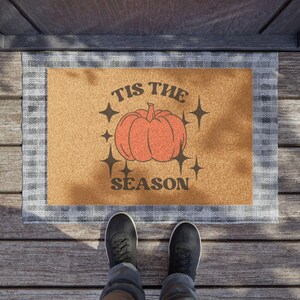 May include: A rectangular doormat with a tan centre and a grey and white plaid border. The centre features an orange pumpkin illustration surrounded by four-pointed stars and the black text TIS THE SEASON printed above and below the pumpkin.