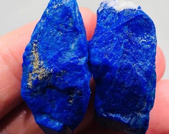 Könnte beinhalten: Zwei rohe, tiefblaue Lapislazuli-Steine mit unregelmäßigen, strukturierten Oberflächen. Der linke Stein weist sichtbare goldene Pyriteinschlüsse auf, während der rechte Stein oben einen weißen Calcitfleck zeigt.