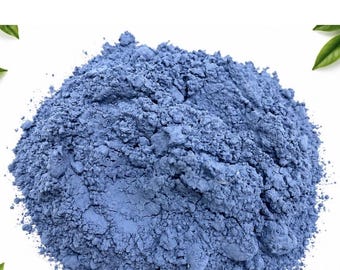 Könnte beinhalten: Ein Haufen aus feinem, pudrigem blauem Pigment mit matter Textur, der kleine Klumpen aufweist. Das Pulver ist auf einem schlichten weißen Hintergrund zentriert, wobei kleine grüne Blätter an den Ecken des Bildes zu sehen sind.