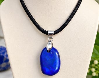 Könnte beinhalten: Eine Halskette mit einem glatten, polierten, tiefblauen ovalen Steinanhänger, der an einer silberfarbenen Metallöse an einem dicken, schwarzen Band hängt. Der Anhänger hat eine glänzende Oberfläche und ist auf einem neutralen Display zentriert.
