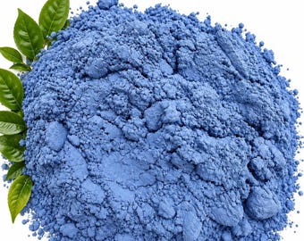 Könnte beinhalten: Ein Haufen feines, leuchtend blaues Pulver mit einer texturierten, körnigen Oberfläche. Mehrere hellgrüne, spitze Blätter sind an der linken Seite des Pulverhaufens angeordnet und bilden einen Kontrast zum tiefen Blau der Substanz.