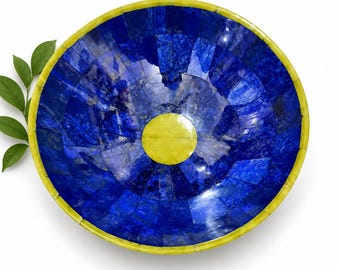 Könnte beinhalten: Eine kreisförmige Schale mit einem Mosaikmuster aus tiefblauen Steinsegmenten in radialer Anordnung. Die Schale hat eine leuchtend gelbe kreisförmige Mitte und einen passenden gelben Rand, mit einem kleinen grünen Blattzweig auf der linken Seite.