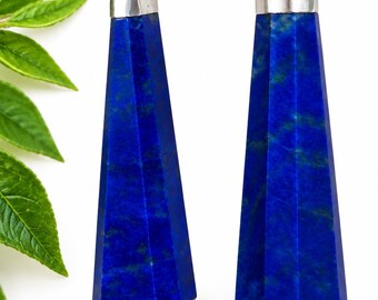 Könnte beinhalten: Zwei facettierte Lapislazuli-Anhänger in tiefem Blau mit subtilen natürlichen Einschlüssen. Jeder Stein ist als langer, spitzer hexagonaler Prismenkörper geschliffen und mit einer polierten silberfarbenen Metallöse versehen.