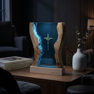 Può includere: Una lampada rettangolare con due bordi in legno naturale che fiancheggiano un centro in resina blu traslucida. Un crocifisso verde è sospeso all'interno della resina. La base è in legno chiaro e reca la scritta RESIN ART VPC.