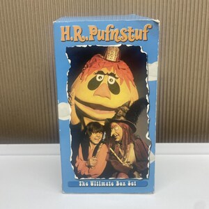 May include: A rectangular H.R. Pufnstuf The Ultimate Box Set product box featuring a light blue background with white cloud accents. The front displays a colorful image of the character Pufnstuf, a child, and a witch with a tall black hat.
