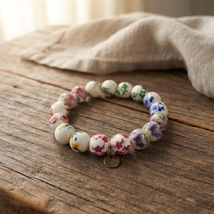 May include: A stretch bracelet composed of round white ceramic beads, each featuring distinct hand-painted floral patterns in shades of red, blue, green, and purple. A small, circular brass-toned metal charm hangs from the bottom of the bracelet.
