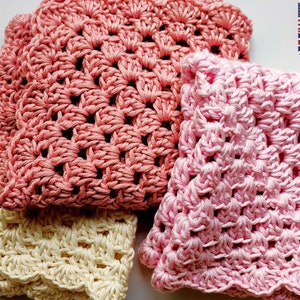 Three hand crocheted washcloths all folded in two and in a random pile, in pink, dusky pink and cream.  Made in a granny square style, with scalloped edging around the outside.  In the top right hand corner are the USA flag and the UK flag.