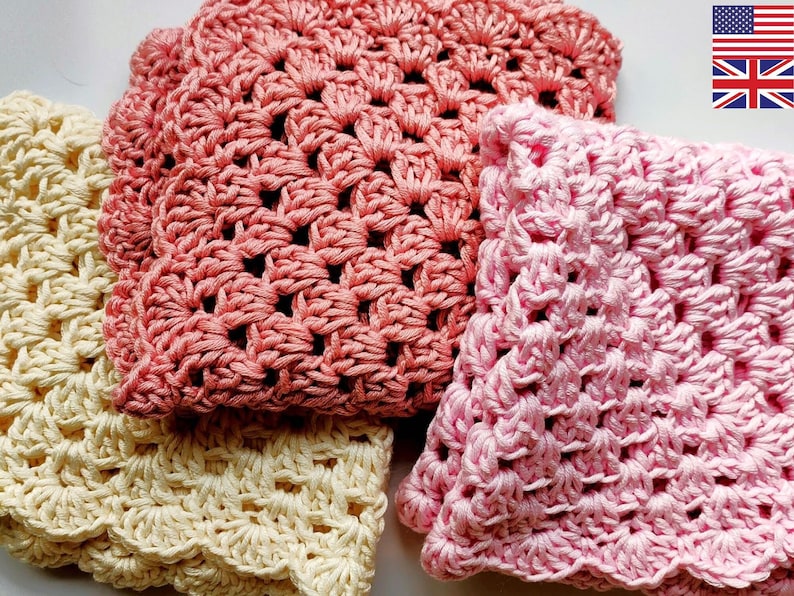 Three hand crocheted washcloths all folded in two and in a random pile, in pink, dusky pink and cream.  Made in a granny square style, with scalloped edging around the outside.  In the top right hand corner are the USA flag and the UK flag.