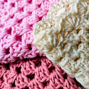 A close up of three crocheted washcloths folded and lying in a display together. One is cream, another dusky pink, the other soft pink.  
The washcloths are a granny square style design, with scalloped edging.
Text on image is PDF Pattern Download.