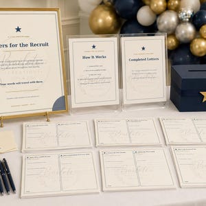 May include: A Letters for the Recruit station featuring a large framed sign, two acrylic instruction signs, a navy blue box with a gold star, cream-coloured stationery cards with printed lines, and a row of navy blue pens on a white tablecloth.