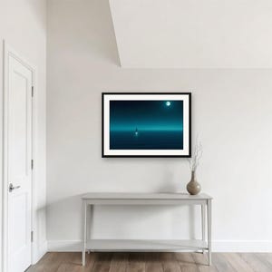 May include: A framed horizontal wall art print featuring a dark teal and blue landscape with a solitary figure standing on a calm, reflective surface under a glowing moon. The artwork is enclosed in a simple black frame and displayed on a white wall.