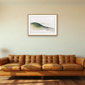 May include: A framed landscape print featuring a watercolour painting of rolling hills in shades of dark green and muted brown, set against a white background. The artwork is enclosed in a simple, light-toned wooden frame with a white mat border.