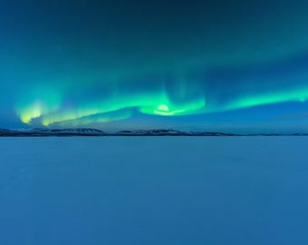 Może przedstawiać: Żywy pokaz zorzy polarnej z wirującymi wstęgami jasnego, zielonego i turkusowego światła rozciągającymi się na ciemnoniebieskim nocnym niebie nad rozległym, płaskim, zaśnieżonym krajobrazem z odległymi, ciemnymi sylwetkami gór na horyzoncie.