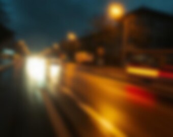 May include: A blurred night scene of a city street featuring bright white headlights from an oncoming vehicle on the left and elongated red and yellow light streaks from moving traffic on the right against a dark, indistinct urban background.