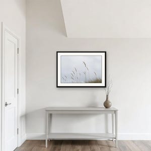 May include: A framed landscape photograph featuring tall, thin blades of grass against a pale, overcast sky. The artwork is displayed in a black frame, mounted on a plain white wall above a light gray console table with a small vase of dried twigs.