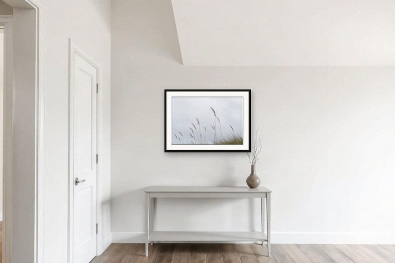 May include: A framed landscape photograph featuring tall, thin blades of grass against a pale, overcast sky. The artwork is displayed in a black frame, mounted on a plain white wall above a light gray console table with a small vase of dried twigs.