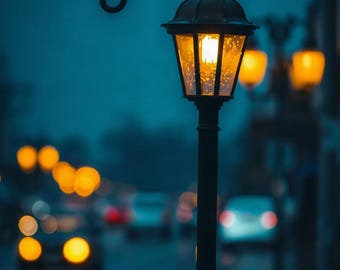 May include: A black metal street lamp with a curved top hook and a glass lantern housing a glowing yellow light. The lamp stands tall against a dark, moody blue background featuring blurred, out-of-focus golden city lights and traffic.