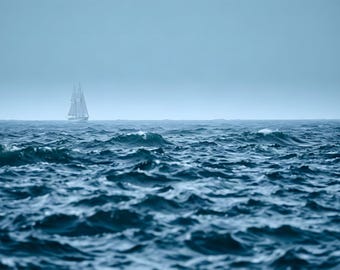 May include: A white two-masted sailing ship navigates across a vast, deep blue ocean with choppy, rolling waves under a hazy, overcast sky. The vessel is positioned in the distance, appearing small against the expansive, dark water.