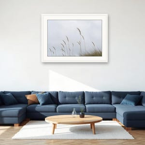 May include: A framed landscape photograph featuring tall, thin blades of beach grass swaying against a pale, overcast sky. The artwork is presented in a clean white frame, showcasing a minimalist nature scene with muted tones and soft lighting.