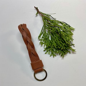 以下が含まれることがあります： 金色のキーリングが付いた、茶色の革製の編み込みキーホルダー。キーホルダーは、緑の葉のついた小枝のある白い背景の上に置かれています。