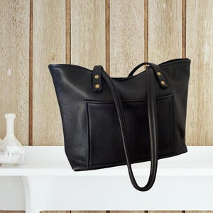 Może przedstawiać: Czarna skórzana torba typu tote z złotymi akcentami i przednią kieszenią. Torba ma dwa długie paski.