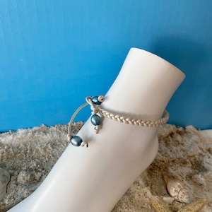 May include: A white braided leather ankle bracelet with blue pearl accents. The bracelet is on a white mannequin foot.