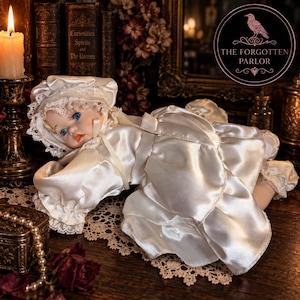 Peut inclure: Une poupée en porcelaine aux yeux bleus et cheveux blonds portant une robe en satin blanc avec bordure en dentelle et un bonnet assorti. La poupée est allongée, avec une finition en tissu brillant, des manches à volants et une jupe ample.