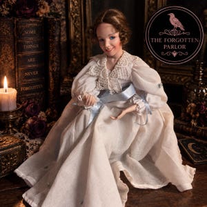 May include: A doll with brown hair and blue eyes wearing a long-sleeved white dress with a lace collar and a light blue ribbon sash. The doll is positioned in front of books and a candle, with a circular logo reading THE FORGOTTEN PARLOR.