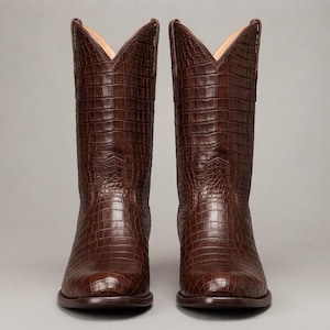 Op de afbeelding: Een paar donkerbruine cowboylaarzen met een textuur van krokodillenleer. De laarzen hebben een hoge schacht met een V-vormige opening aan de bovenkant, een ronde neus en een zichtbare hak. Het buitenoppervlak vertoont een consistent rasterpatroon.