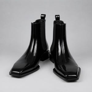 Puede incluir: Un par de botines negros con acabado de charol brillante, que presentan una punta cuadrada, un tacón bajo de bloque y paneles laterales elásticos. Cada bota incluye una lengüeta vertical en la parte trasera y una costura central visible.