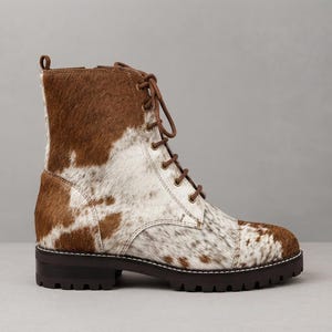 Pode incluir: Uma bota de cano curto com atacadores, apresentando um padrão de pele de vaca em castanho e branco com acabamento texturizado de pelo. A bota inclui atacadores castanhos, ilhós em latão, biqueira reforçada e sola de borracha grossa e escura.