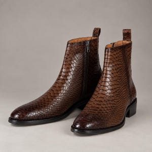 Peut inclure: Une paire de bottines avec un motif texturé en peau de serpent marron. Les bottines présentent un bout pointu, une fermeture éclair latérale avec une tirette argentée, une languette au talon et un petit talon bloc avec une semelle foncée.