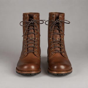 Op de afbeelding: Een paar halfhoge veterschoenen van rijk bruin leer met een textuur. Deze laarzen zijn voorzien van donkerbruine veters door messingkleurige metalen oogjes, een ronde neus en een dikke, donkerbruine profielzool voor extra grip.