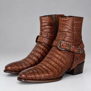 Puede incluir: Un par de botines marrones con un patrón de textura de piel de cocodrilo. Cada bota incluye una cremallera lateral, una correa decorativa en el tobillo y una hebilla de metal plateado con grabados intrincados. Tacón de bloque bajo.