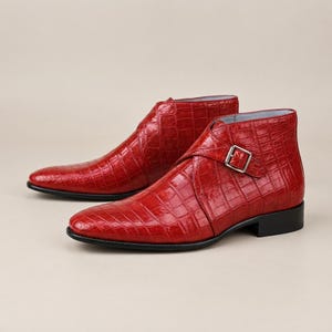 Puede incluir: Un par de botines de color rojo vibrante con una textura brillante de estampado de cocodrilo. Cada bota tiene una punta afilada, un tacón bajo negro y una correa lateral única asegurada con una hebilla de metal plateado en el tobillo.