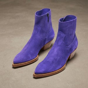 May include: A pair of vibrant purple suede ankle boots featuring a pointed toe, a side zip closure, and a stacked wooden heel. The boots have a clean, unadorned upper with subtle stitching details around the ankle and a natural wood-toned sole.