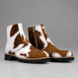 Peut inclure: Une paire de bottines avec un motif de vache marron et blanc et une finition texturée en poils. Les bottines sont dotées d'une fermeture éclair latérale, d'un bout arrondi et d'une épaisse semelle crantée noire avec une couture apparente.
