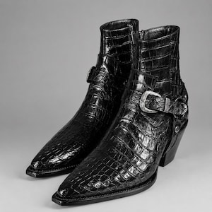Könnte beinhalten: Ein Paar schwarze Stiefeletten mit glänzender Krokodilprägung, spitzer Zehenpartie und einem seitlichen Riemen mit silberfarbener Metallschnalle. Die Stiefel verfügen über einen Blockabsatz und einen seitlichen Reißverschluss.