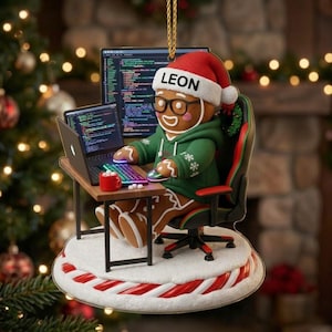 May include: A gingerbread person ornament wearing a green hoodie, black glasses, and a red Santa hat with the name LEON printed on the brim. The figure sits in a gaming chair at a desk with a laptop displaying code, a mouse, and a red mug.
