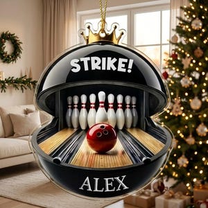 May include: A black, circular, flat acrylic ornament featuring a gold crown at the top. The center displays a bowling alley scene with white pins and a red bowling ball. The text STRIKE! is printed in white above, and the name ALEX is printed below.