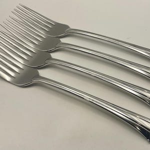 May include: A set of four matching stainless steel dinner forks with a polished silver finish. Each fork features a four-tine head and a handle that widens toward the base, decorated with three subtle, vertical grooved lines near the end.