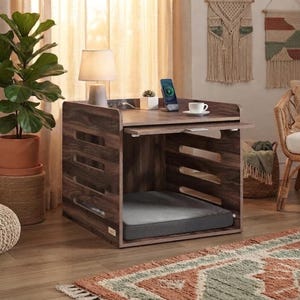 May include: A dark wood pet crate end table featuring a flat top surface, a pull-out tray, and horizontal side slats. The interior contains a removable gray rectangular cushion. The top surface holds a lamp, a small succulent, and a phone.