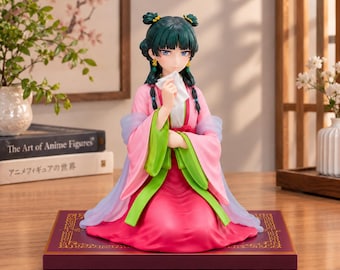 Puede incluir: Una figura coleccionable de anime con cabello verde azulado oscuro recogido en moños, vistiendo una túnica tradicional de color rosa y verde. La figura sostiene un pequeño paño blanco frente a su rostro y está arrodillada sobre una base morada.