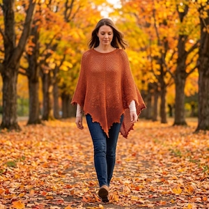 May include: A rust-colored, open-knit poncho featuring a textured, diamond-patterned weave with a pointed hemline. The garment is worn over a long-sleeved, light-colored base layer and paired with dark blue denim jeans, creating a layered look.
