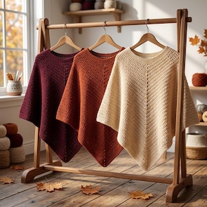 May include: Three crocheted ponchos hanging on a wooden rack. The ponchos are in deep burgundy, burnt orange, and cream colors. Each garment features a textured stitch pattern with a central vertical seam and a pointed hemline at the bottom.