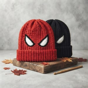 Könnte beinhalten: Zwei gerippte Strickmützen, eine rote und eine schwarze, die jeweils mit einem Paar weißer, schwarz umrandeter Augen auf der Vorderseite versehen sind. Die Mützen liegen auf einem Holzblock mit Herbstblättern und einer Häkelnadel aus Holz.
