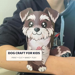 May include: A paper craft dog figure featuring brown and white fur, large black eyes, and a pink yarn ball. The text on the craft reads: DOG CRAFT FOR KIDS, PRINT • CUT • MAKE • PLAY. The figure is designed to wrap around an arm.