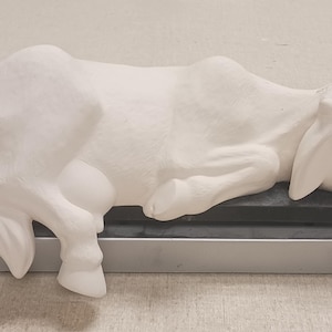 May include: A white ceramic cow figurine in a lying position, resting on a digital scale. The cow has a smooth, unpainted surface with visible sculpted details including ears, legs, and a tail. The scale is silver and black with a digital display.