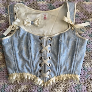 May include: A light blue corset top featuring a white star and swirl pattern, a front white ribbon lace-up closure with metal grommets, cream-colored ruffled trim at the hem, and white ribbon shoulder ties. A small white bow is attached to the chest.